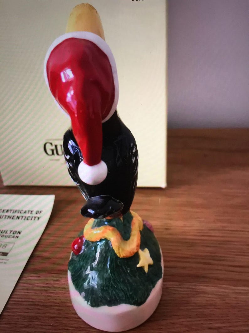 May include: A black bird figurine wearing a red Santa hat and a yellow scarf, standing on a green Christmas tree base with a pink bottom.
