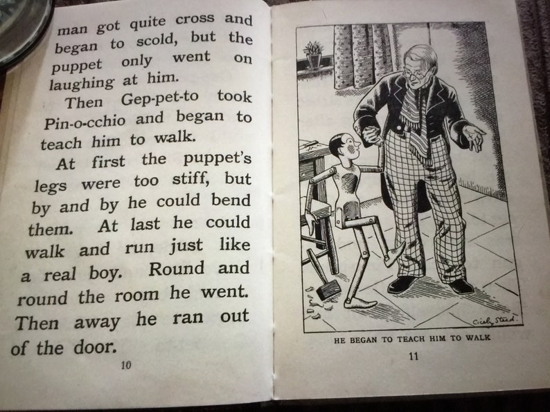 May include: An open book with illustrations and text. The right page shows a black and white drawing of a man teaching a wooden puppet to walk. The text reads "HE BEGAN TO TEACH HIM TO WALK".
