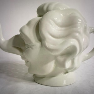 Puede incluir: Una tetera de cerámica blanca con un diseño único. La tetera tiene forma de rostro, con una trompa de elefante como pico y un asa lateral. La cara tiene rasgos detallados y un diseño de pelo rizado en la parte superior.