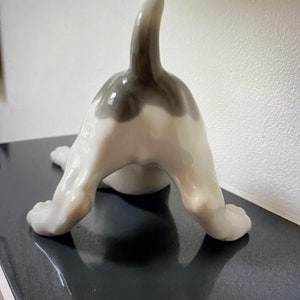 May include: A white porcelain figurine of a dog with a black tail and black markings on its back. The dog is in a playful pose, with its hind legs raised and its head tilted back.