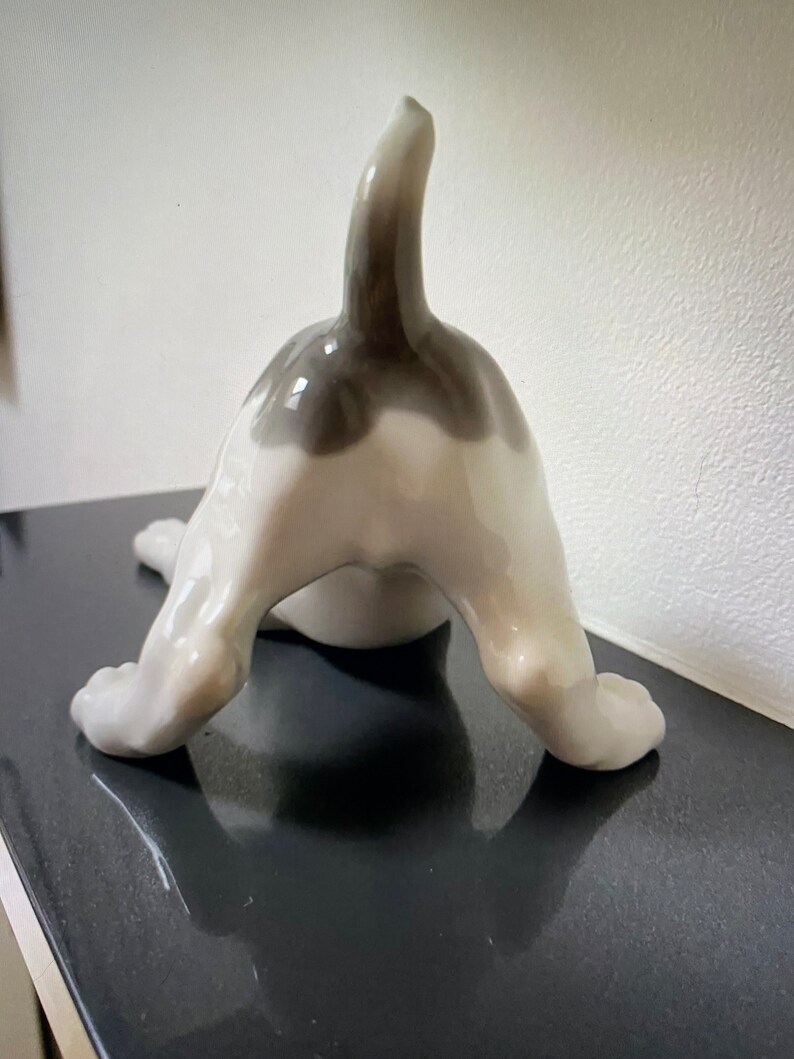 May include: A white porcelain figurine of a dog with a black tail and black markings on its back. The dog is in a playful pose, with its hind legs raised and its head tilted back.