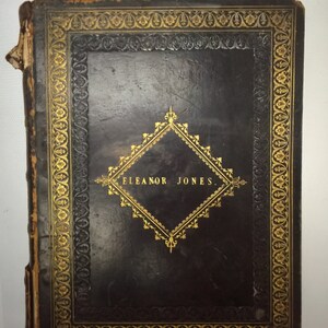 May include: A black leather-bound book with gold-colored decorative borders and a diamond-shaped design in the center. The text "ELEANOR JONES" is inscribed in the center of the diamond.