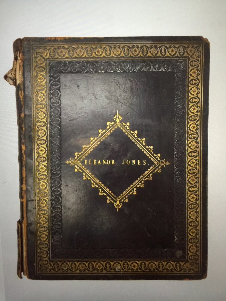 May include: A black leather-bound book with gold-colored decorative borders and a diamond-shaped design in the center. The text "ELEANOR JONES" is inscribed in the center of the diamond.