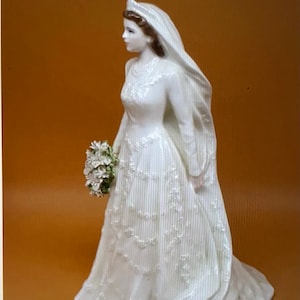 May include: A white porcelain figurine of a bride wearing a long, flowing gown with a veil and holding a bouquet of white flowers.