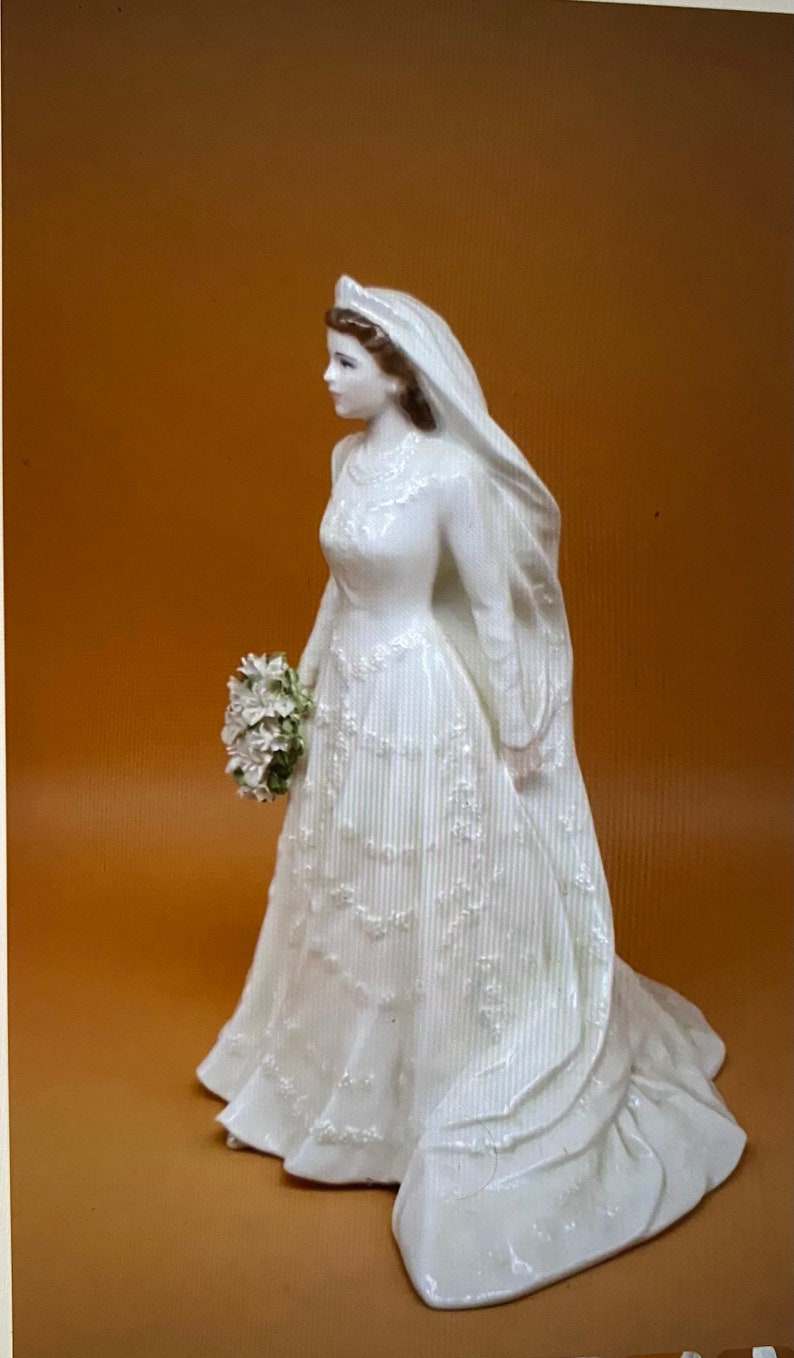May include: A white porcelain figurine of a bride wearing a long, flowing gown with a veil and holding a bouquet of white flowers.