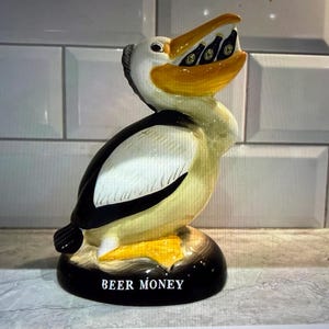 May include: A ceramic pelican-shaped bank with a yellow beak and a black and white body. The pelican's beak holds several small beer bottles. The base of the bank is black with the words "BEER MONEY" in white.