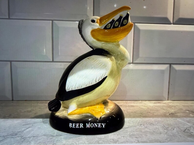 May include: A ceramic pelican-shaped bank with a yellow beak and a black and white body. The pelican's beak holds several small beer bottles. The base of the bank is black with the words "BEER MONEY" in white.