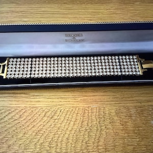 May include: A wide gold-tone bracelet featuring five rows of clear round rhinestones, presented in a dark blue rectangular hinged display box with the text Brooks & Bentley printed in gold on the interior white satin lining of the lid.