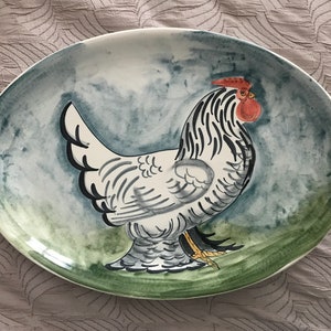 Può includere: Un piatto da portata ovale in ceramica con un design dipinto a mano di un gallo bianco con strisce nere e una cresta rossa. Il gallo è in piedi su un campo verde con uno sfondo acquerellato blu e verde.