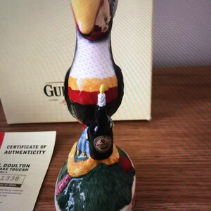 May include: A ceramic figurine of a toucan holding a bottle of Guinness beer. The toucan is wearing a red, white, and yellow hat and has a blue and white scarf. The figurine is standing on a green base with a brown and yellow snake wrapped around it. The base is decorated with flowers and leaves. The figurine is part of a collection of Doulton Christmas Toucans.