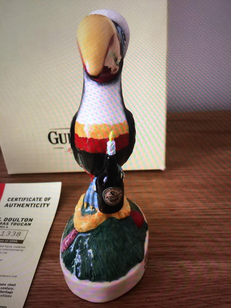 May include: A ceramic figurine of a toucan holding a bottle of Guinness beer. The toucan is wearing a red, white, and yellow hat and has a blue and white scarf. The figurine is standing on a green base with a brown and yellow snake wrapped around it. The base is decorated with flowers and leaves. The figurine is part of a collection of Doulton Christmas Toucans.