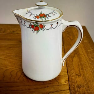 May include: White ceramic jug with a lid, gold trim, and a handle. The jug is decorated with a floral design of red and green fruit. It is likely used for serving beverages, with a capacity of approximately 1.5 liters.