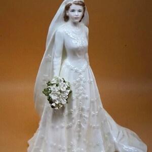 May include: A white porcelain figurine of a bride wearing a white gown with a veil and holding a bouquet of white flowers.