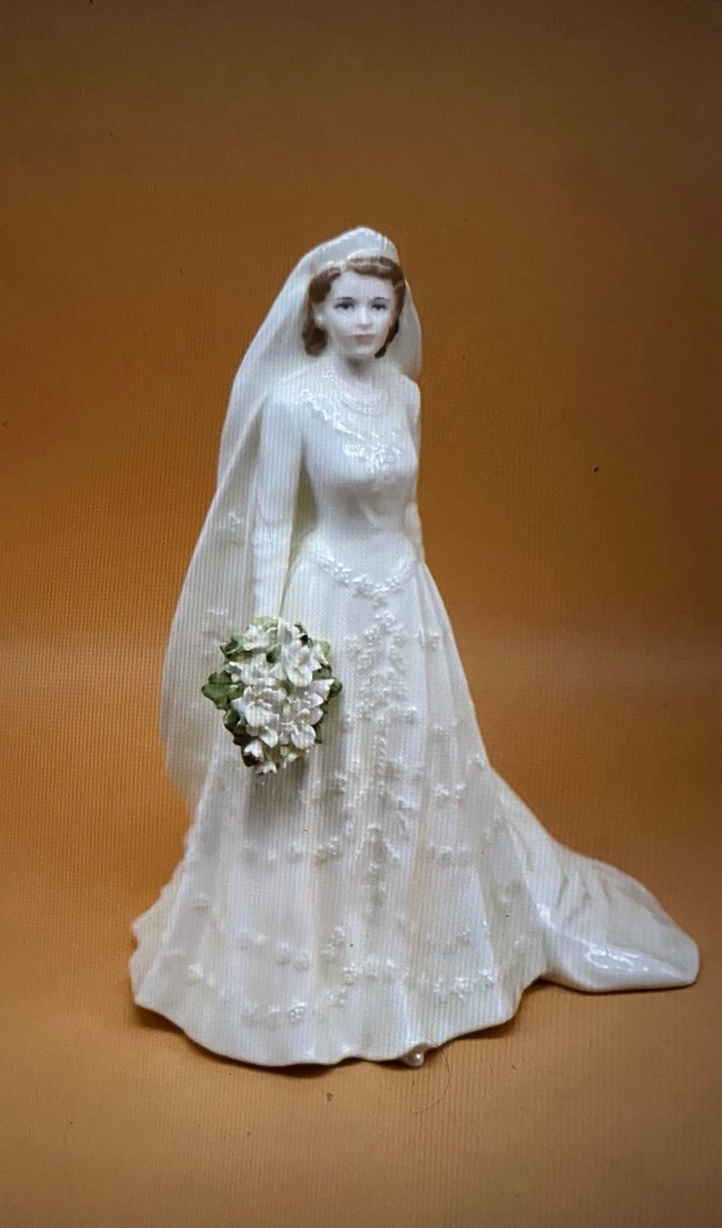 May include: A white porcelain figurine of a bride wearing a white gown with a veil and holding a bouquet of white flowers.