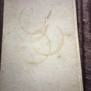 May include: A vintage book with a light beige cover. The cover has several large, faded brown stains in a circular pattern. The book's edges are slightly worn, suggesting age and use. The overall aesthetic is rustic and aged.