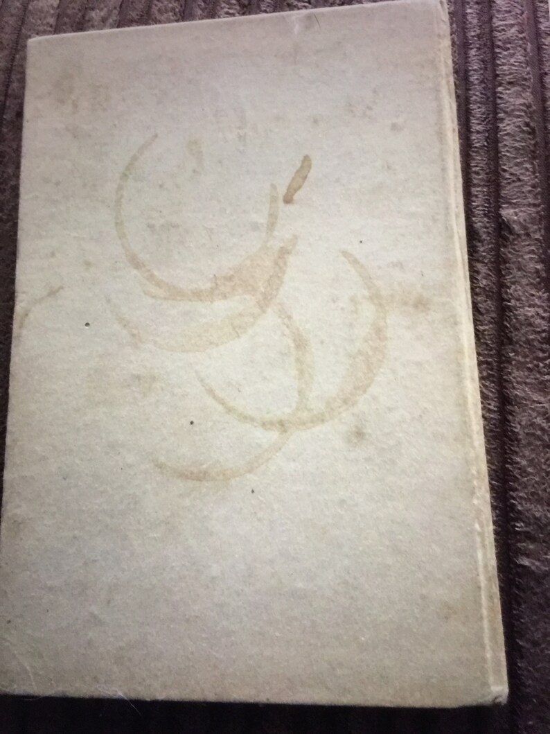 May include: A vintage book with a light beige cover. The cover has several large, faded brown stains in a circular pattern. The book's edges are slightly worn, suggesting age and use. The overall aesthetic is rustic and aged.