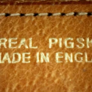 Puede incluir: Primer plano de una superficie de cuero marr&oacute;n con el texto "REAL PIGSKIN MADE IN ENGLAND" en relieve en blanco.
