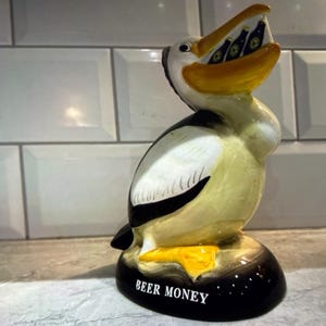 May include: A ceramic pelican figurine with a yellow beak and feet, holding miniature beer bottles in its mouth. The bird has black and white markings and the base reads "BEER MONEY".