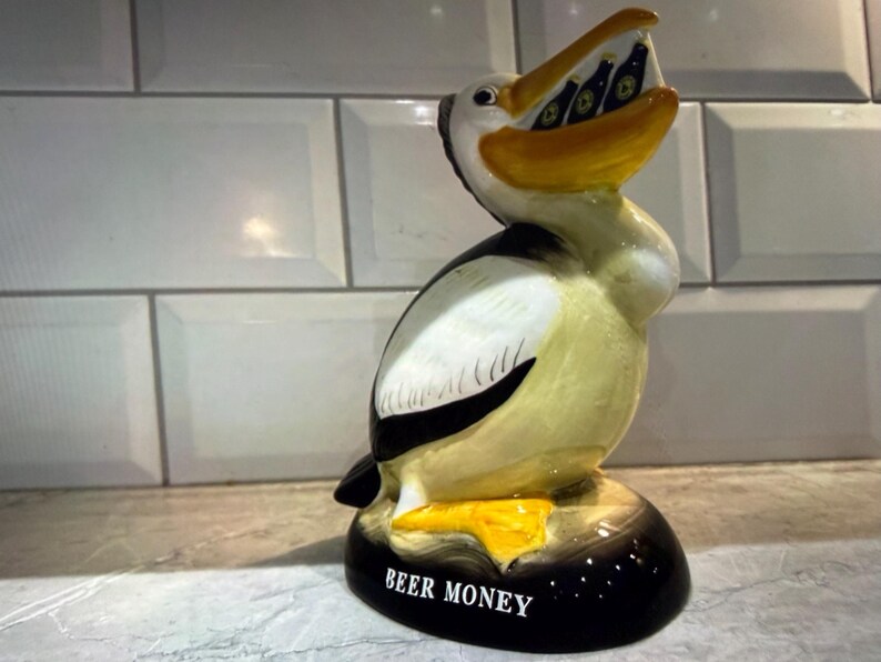 May include: A ceramic pelican figurine with a yellow beak and feet, holding miniature beer bottles in its mouth. The bird has black and white markings and the base reads "BEER MONEY".