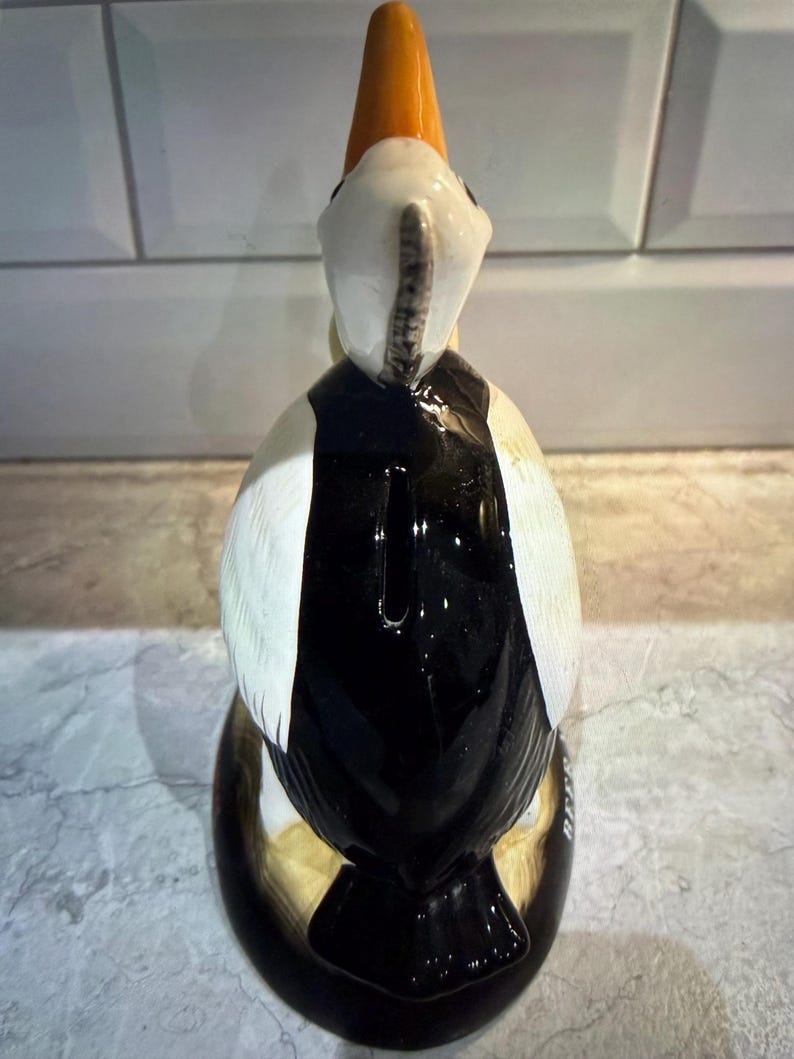 May include: A ceramic puffin figurine with a black and white body, white wings, and an orange beak. The bird is standing upright, with a glossy finish. The figurine is a decorative item.