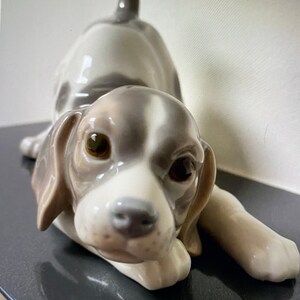 May include: A porcelain figurine of a white and brown puppy with floppy ears, lying down with its head resting on its paws. The puppy has brown eyes and a black nose.