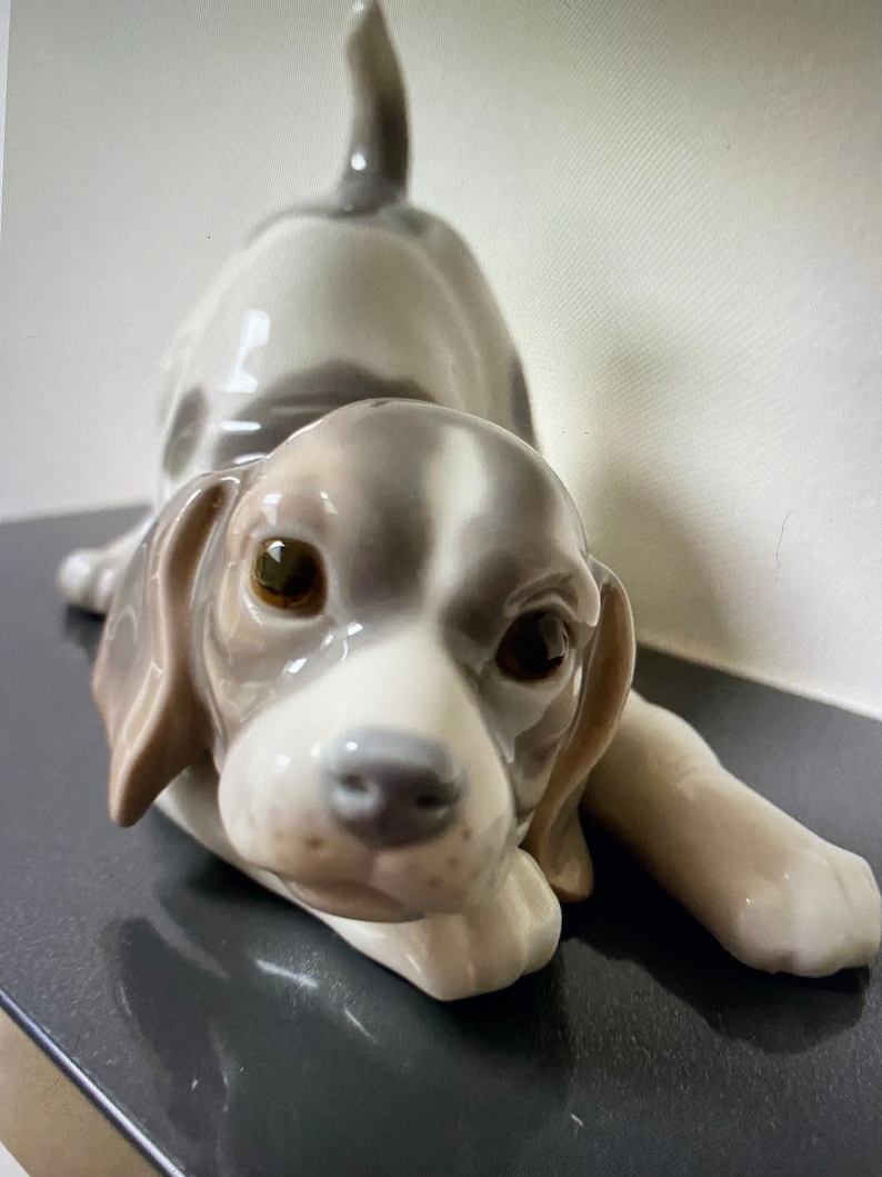 May include: A porcelain figurine of a white and brown puppy with floppy ears, lying down with its head resting on its paws. The puppy has brown eyes and a black nose.