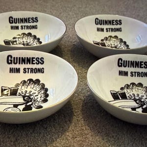 May include: Four white ceramic bowls with a gold rim. Each bowl has the text "Guinness Him Strong" in black, along with a black and white graphic of a toucan and a pint of Guinness.