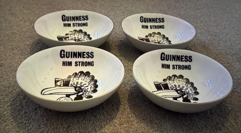 May include: Four white ceramic bowls with a gold rim. Each bowl has the text "Guinness Him Strong" in black, along with a black and white graphic of a toucan and a pint of Guinness.