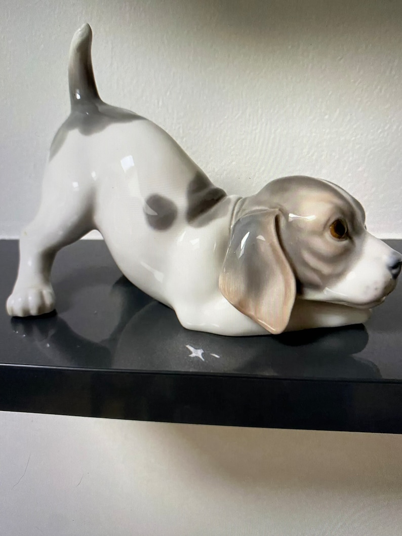 May include: A porcelain figurine of a white and gray puppy with brown eyes, lying on its belly with its tail curled up. The puppy is looking to the right.