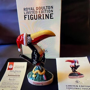 May include: A Royal Doulton limited edition Christmas toucan figurine. The toucan is wearing a Santa hat and is perched on a green tree with a snake. The base of the figurine reads "Guinness is good for Yule".