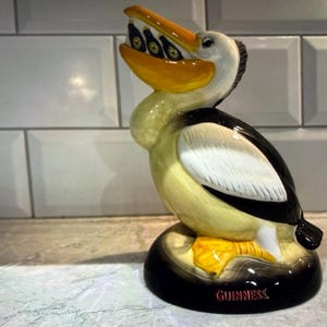 May include: A ceramic pelican figurine with a yellow beak and feet, black and white wings, and a cream-colored body. The pelican holds several small bottles in its beak. The base is black with the word "GUINNESS" in red.