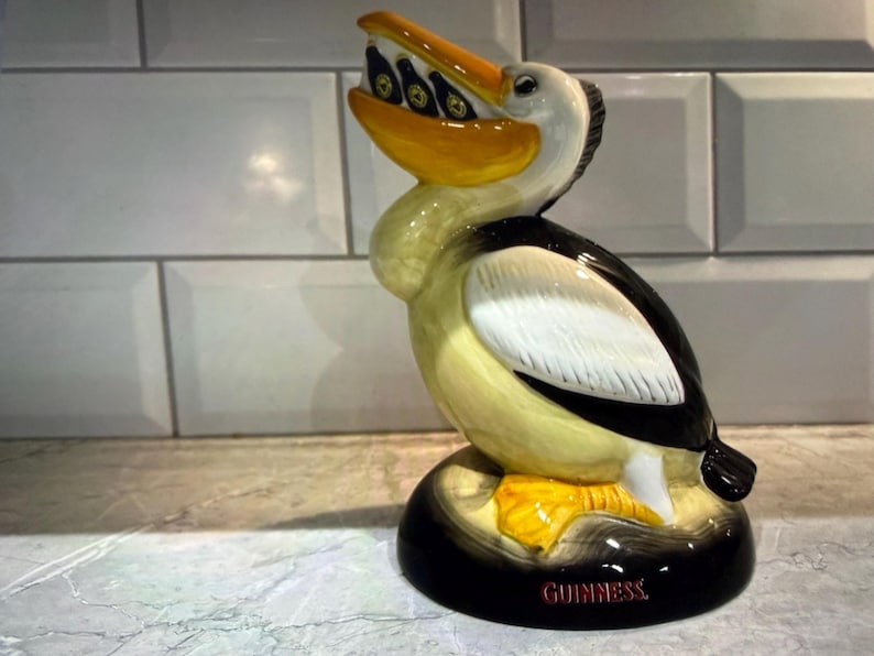 May include: A ceramic pelican figurine with a yellow beak and feet, black and white wings, and a cream-colored body. The pelican holds several small bottles in its beak. The base is black with the word "GUINNESS" in red.