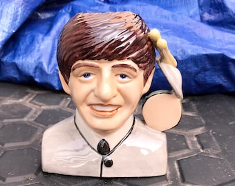 May include: A ceramic figurine of a man with brown hair, wearing a gray jacket and a black tie. The figurine is holding a drum and drumsticks in his right hand.