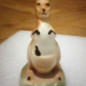 K&ouml;nnte beinhalten: Keramik-K&auml;nguru-Figur mit einem beige-cremefarbenen K&ouml;rper, schwarzen Akzenten und einem gr&uuml;nen Sockel. Das K&auml;nguru hat aufrechte Ohren und ein detailliertes Gesicht. Die Figur ist ein dekoratives Objekt.