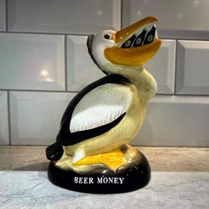 May include: A ceramic pelican figurine with a black base and the words "BEER MONEY" in white. The pelican has a yellow beak and is holding miniature beer bottles. The bird's body is white, black, and tan.
