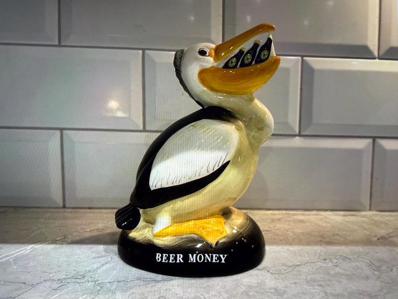 May include: A ceramic pelican figurine with a black base and the words "BEER MONEY" in white. The pelican has a yellow beak and is holding miniature beer bottles. The bird's body is white, black, and tan.
