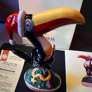 May include: A ceramic toucan figurine wearing a red Santa hat, perched on a green base with a gold star and a snake. The base reads "Guinness and the Toucan".