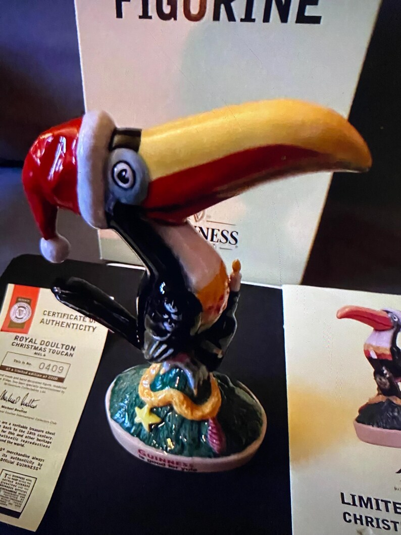 May include: A ceramic toucan figurine wearing a red Santa hat, perched on a green base with a gold star and a snake. The base reads "Guinness and the Toucan".
