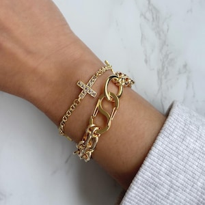 May include: Two gold bracelets on a wrist. One is a delicate chain with a cross pendant, the other is a chunky chain link style. The bracelets are displayed on a light background.