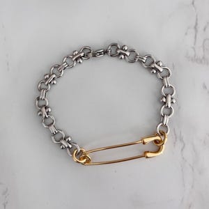 Può includere: Bracciale a catena color argento con maglie decorative e chiusura a spilla da balia dorata. Il bracciale è esposto su una superficie di marmo bianco e grigio. Un gioiello unico.