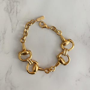 May include: A gold bracelet with a horse bit design. The bracelet features a series of connected horse bit-shaped links, with a chain and clasp closure. The jewellery is displayed on a white and grey marble surface.