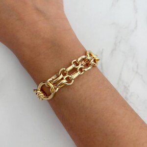 May include: A gold-colored chain link bracelet with a double-strand design. The bracelet is made of a shiny, polished metal and features a large clasp. The bracelet is shown on a person's wrist.