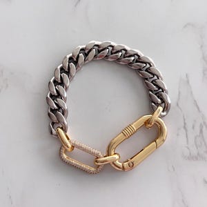 May include: A silver and gold chain bracelet. The bracelet features a thick, silver chain that connects to a gold clasp. A smaller gold link with clear stones connects the chain to the clasp.