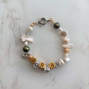 Könnte beinhalten: Ein Perl- und Goldperlenarmband mit dem Namen "JORDAN" in silbernen und goldenen Buchstabenperlen.
