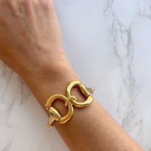 Op de afbeelding: Een gouden armband met een groot, in elkaar grijpend, dubbel-lus ontwerp. De armband zit om een pols.