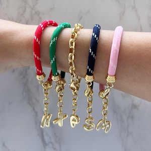 May include: A selection of bracelets in various colours. The bracelets have red, green, navy, and pink rope-like bands, and a gold chain. Each bracelet has a gold charm with the letters "HOPE" and "SM".