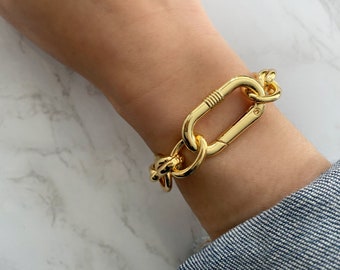 May include: A gold-colored bracelet featuring a large, rectangular clasp and chain links. The bracelet is worn on a wrist, showcasing its shiny, metallic finish. The design is bold and modern, suitable for everyday wear.