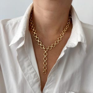 Könnte beinhalten: Eine goldfarbene Y-förmige Halskette, die aus einer Reihe miteinander verbundener kreisförmiger Glieder besteht. Die Kette bildet eine durchgehende Schlaufe um den Hals mit einem vertikalen Anhänger, der von der Mitte herabhängt.