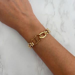 May include: Gold-toned chain bracelet featuring a large clasp and chunky links. The bracelet is made of a shiny, polished metal and is worn on a wrist. The design is bold and eye-catching, suitable for adding a touch of glamour to any outfit.