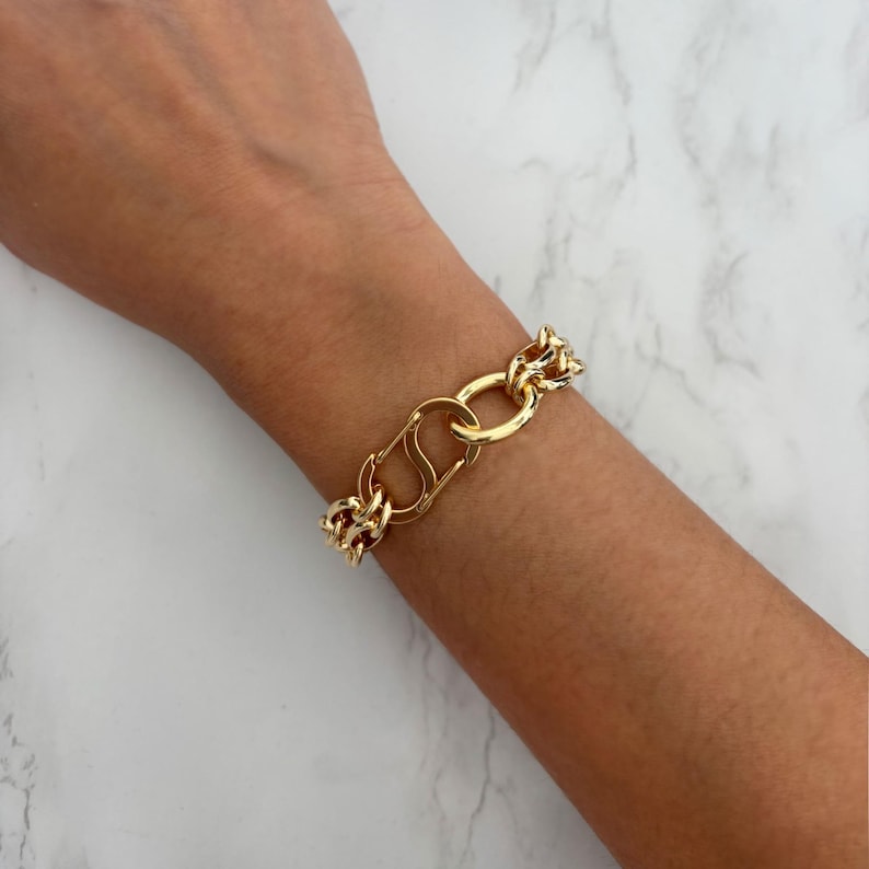 May include: Gold-toned chain bracelet featuring a large clasp and chunky links. The bracelet is made of a shiny, polished metal and is worn on a wrist. The design is bold and eye-catching, suitable for adding a touch of glamour to any outfit.